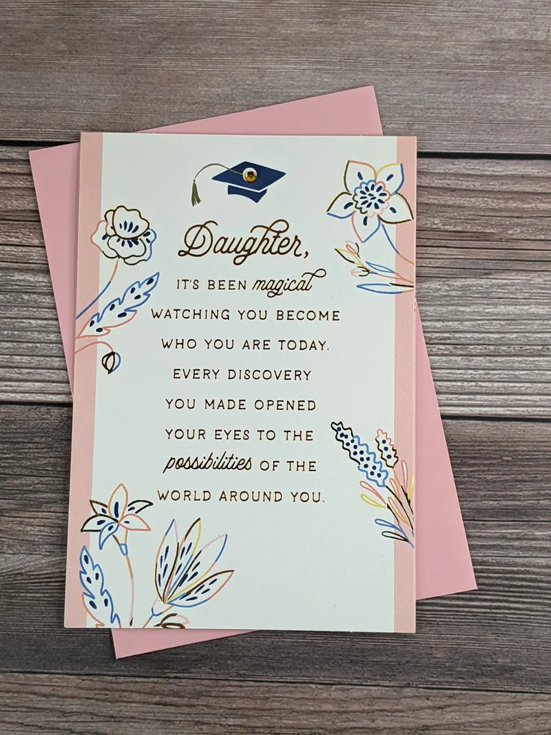 A graduation card for a daughter, featuring a blue grad cap with a yellow gem, a flower design with pink accents, and a pink envelope.