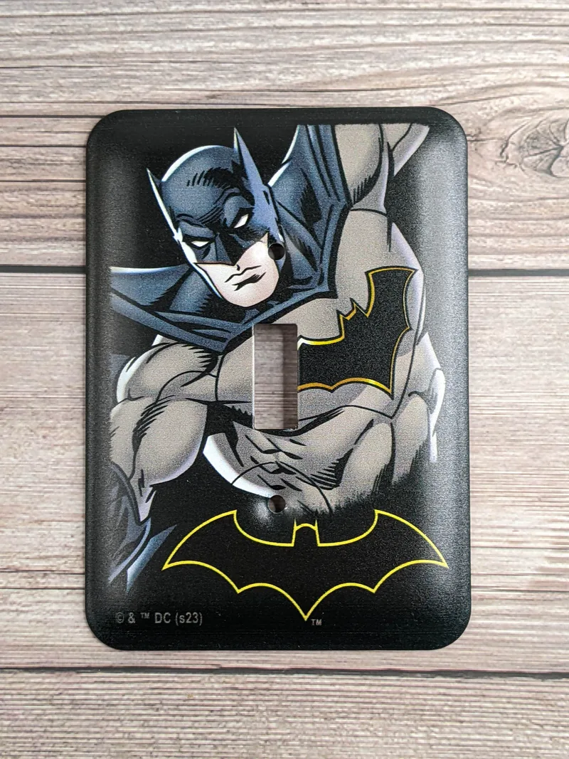 A Justice League DC Comics light switch plate cover that is black with an image of Batman and a Batman symbol.