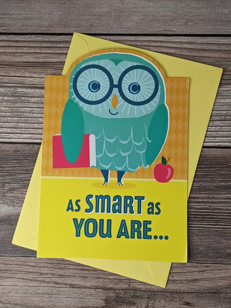 A yellow greeting card with an image on the cover of a blue owl holding a schoolbook next to a red apple.