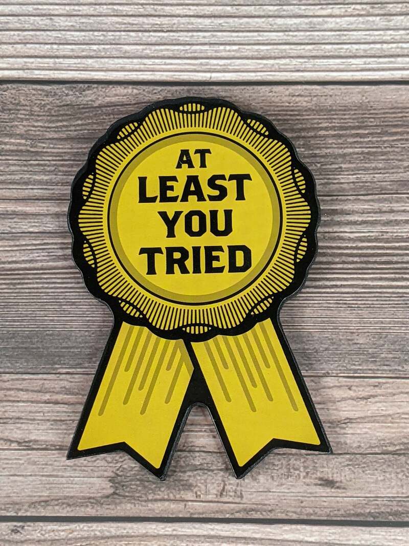 A yellow magnet shaped like a ribbon award with black lettering of the words, "at least you tried."